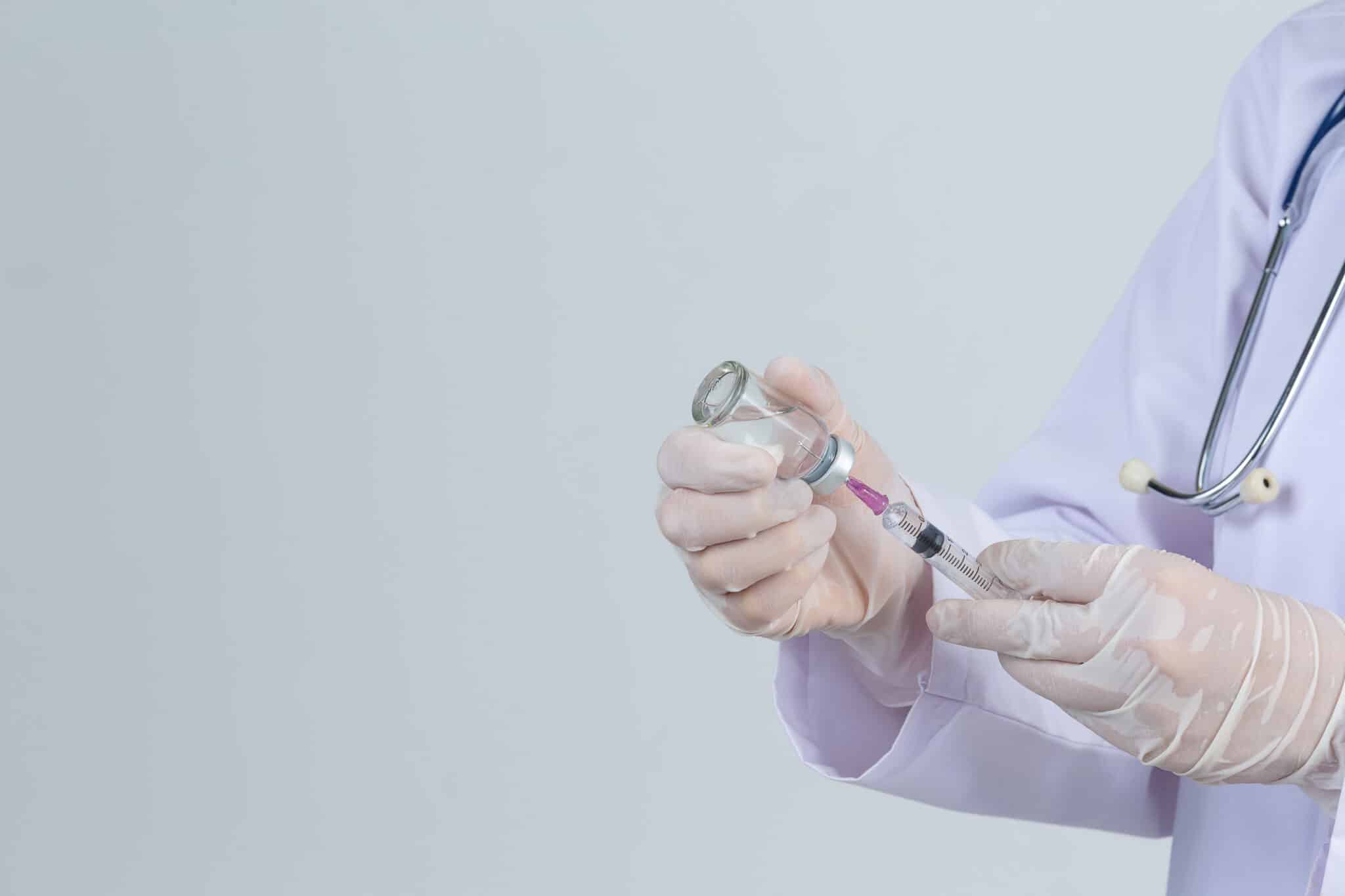 Young doctor is  holding  hypodermic syringe with  vaccine vial  rubber gloves on gray background.